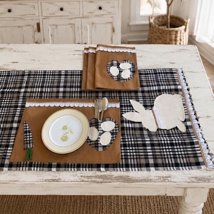 May include: A brown placemat and table runner set featuring a black and white plaid pattern, white lace trim, and fabric appliqués of a rabbit, a carrot, and hearts with white ovals. A round white plate with a floral design sits on the placemat.