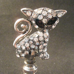 May include: A silver lamp finial with a rhinestone-covered cat design. The cat has black eyes and a curled tail.