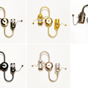 May include: A collection of five double-socket light fixtures in various metallic finishes including brass, gold, bronze, black, and silver. Each fixture features a central circular wall mount with two curved arms, each ending in a socket with a pull chain.