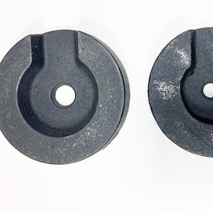 May include: Two black metal targets with a circular shape and a cutout in the center. The targets have a textured surface and a small hole in the center.