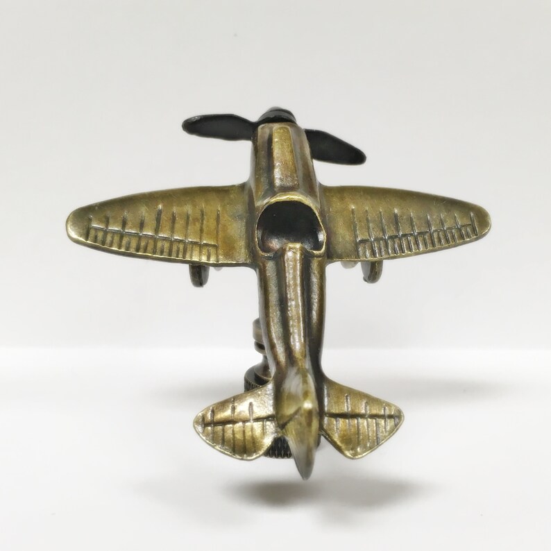 Puede incluir: Una figurita de avi&oacute;n de metal de color dorado con palas de h&eacute;lice negras. El avi&oacute;n est&aacute; detallado con l&iacute;neas y tiene un acabado ligeramente desgastado.
