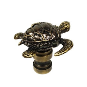 May include: A brass lamp finial featuring a detailed sea turtle sculpture. The turtle is facing right and has a textured shell. The finial is designed to be used on a lamp shade.