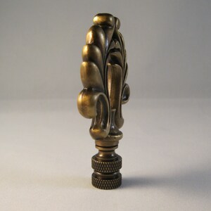 May include: A brass lamp finial with a decorative, ornate design. The finial has a fluted, leaf-like pattern and a rounded top.