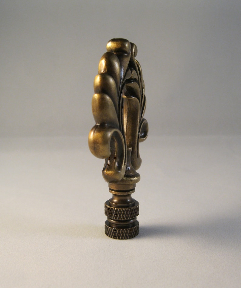 May include: A brass lamp finial with a decorative, ornate design. The finial has a fluted, leaf-like pattern and a rounded top.