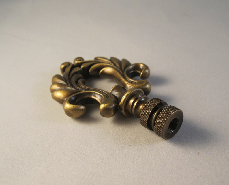 May include: A decorative brass finial with a detailed floral design. The finial has a threaded end for attaching to a lampshade or other object.