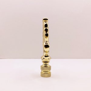 May include: Gold metal lamp finial with a decorative design. The finial has a round base and a pointed top.