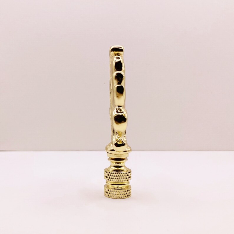 May include: Gold metal lamp finial with a decorative design. The finial has a round base and a pointed top.