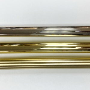 May include: Three metal rods in different finishes: dark bronze, polished brass, and gold. Each rod is cylindrical with a cap on each end. The rods are displayed horizontally against a white background, showcasing their reflective surfaces and varying hues.