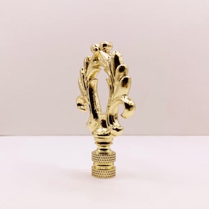 May include: A gold-toned lamp finial with a decorative, ornate design featuring a stylized leaf pattern.
