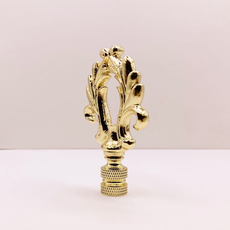 May include: A gold-toned lamp finial with a decorative, ornate design featuring a stylized leaf pattern.