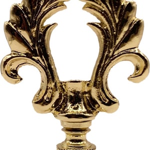 May include: Gold metal lamp finial with a decorative, ornate design. The finial has a leaf-like pattern and a round opening in the center.
