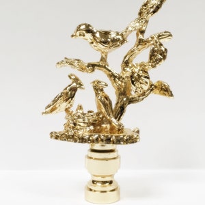 May include: Gold metal lamp finial with a detailed design of birds in a tree. The finial is a decorative piece for a lamp.