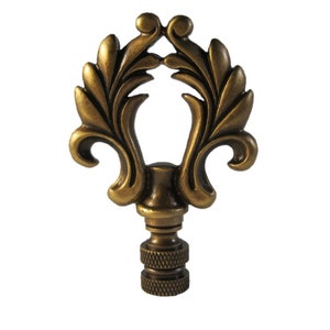 May include: A gold-toned metal lamp finial with a decorative, ornate design featuring a stylized leaf motif. The finial has a threaded base for attaching to a lampshade.