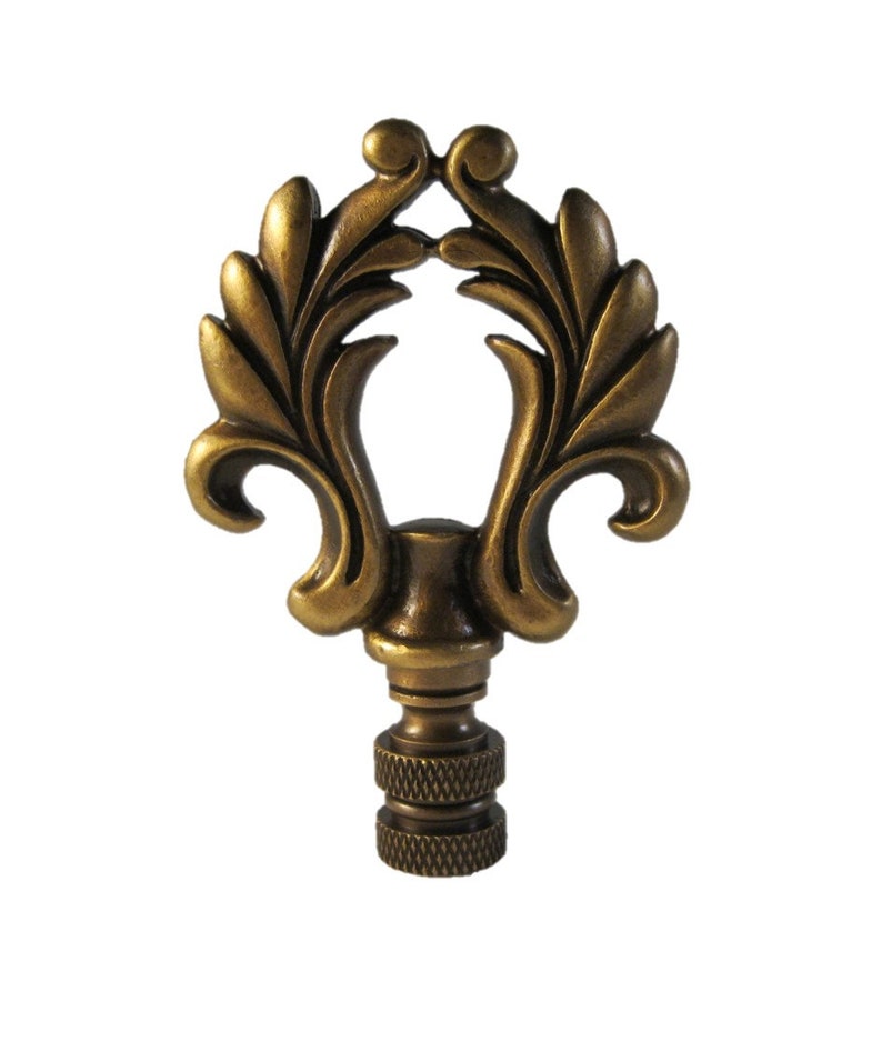 May include: A gold-toned metal lamp finial with a decorative, ornate design featuring a stylized leaf motif. The finial has a threaded base for attaching to a lampshade.