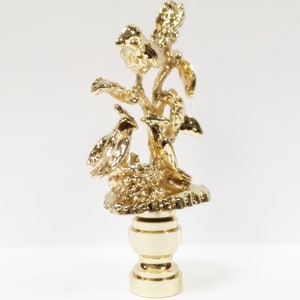May include: Gold lamp finial with a detailed design of birds perched on a branch with leaves. The finial is made of metal and has a smooth, polished finish.