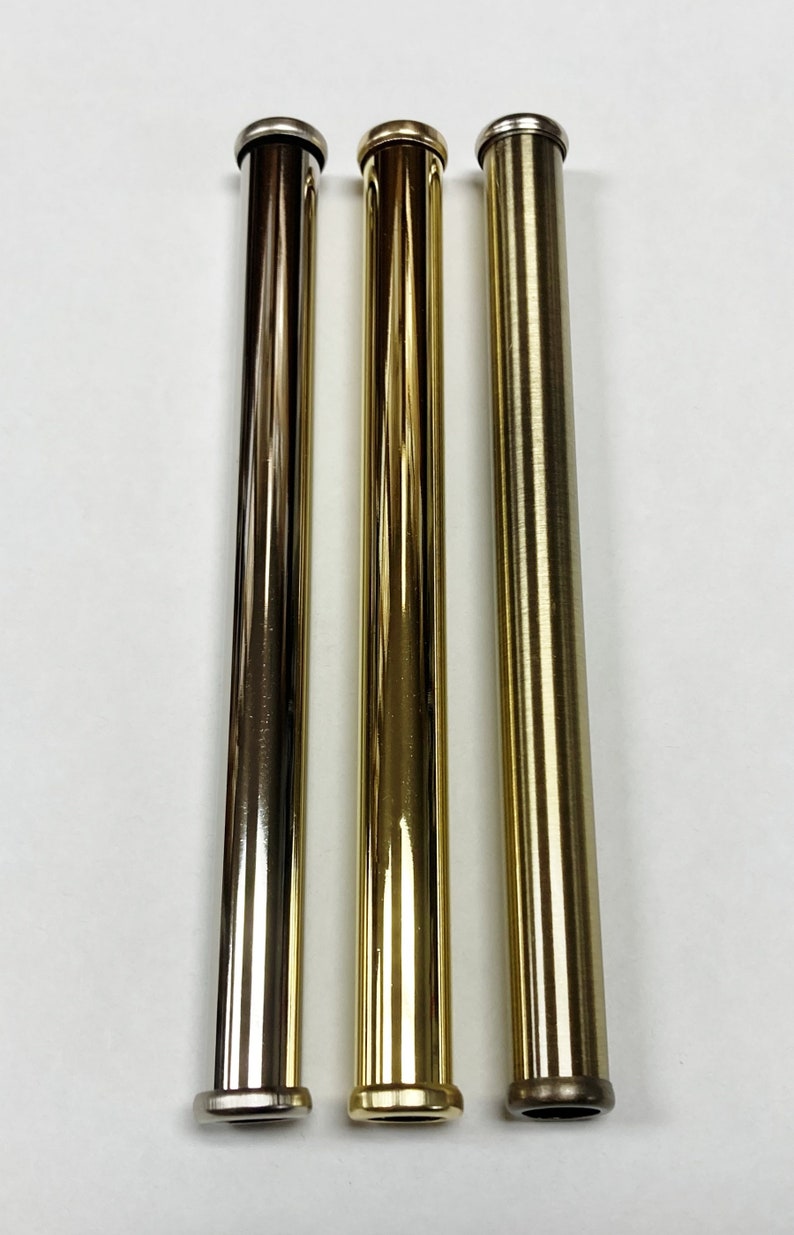 May include: Three metallic, cylindrical tubes in varying shades of gold and bronze. Each tube has a rounded top and bottom, with a polished finish. The tubes are arranged vertically against a white background.