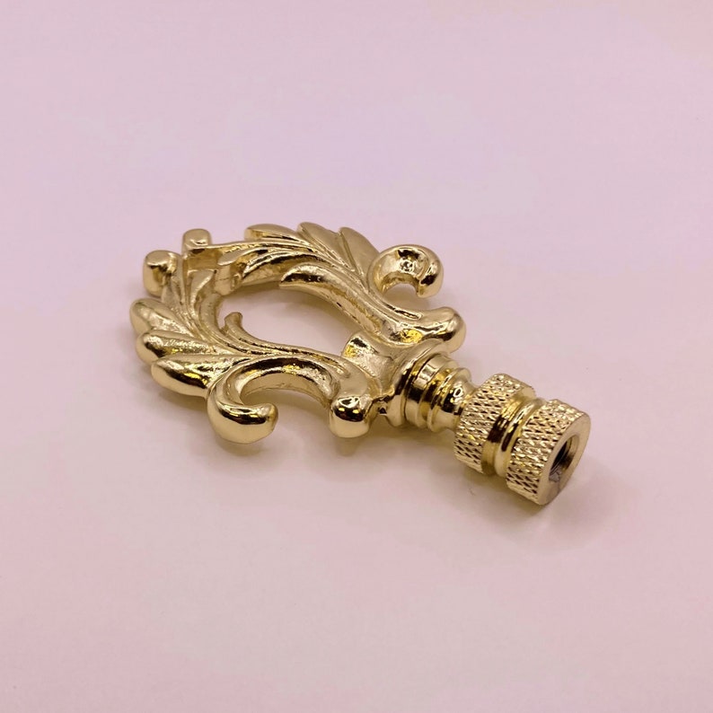 May include: Gold metal lamp finial with a decorative, ornate design. The finial has a threaded base for attaching to a lamp.