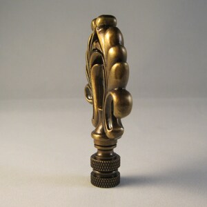 May include: A gold-toned metal lamp finial with a decorative, ornate design. The finial has a fluted, scroll-like shape with a rounded top.