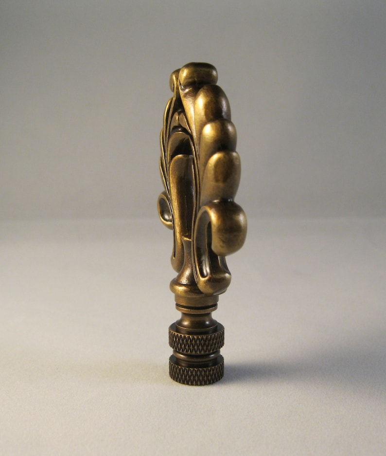May include: A gold-toned metal lamp finial with a decorative, ornate design. The finial has a fluted, scroll-like shape with a rounded top.