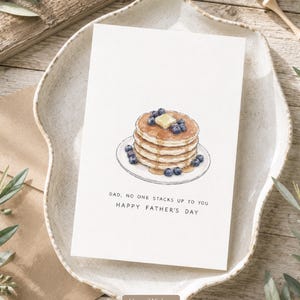 May include: A white greeting card featuring a watercolor illustration of a stack of pancakes topped with blueberries and a pat of butter on a plate. The text reads: DAD, NO ONE STACKS UP TO YOU. HAPPY FATHER'S DAY. The card is rectangular.