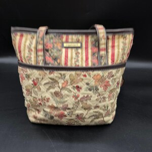 May include: A tote bag featuring a beige base with a woven floral and leaf pattern in shades of red, green, and brown. The bag has dark brown trim, two shoulder straps, and a rectangular metal plaque on the front with the text "TAPESTRY" in gold.