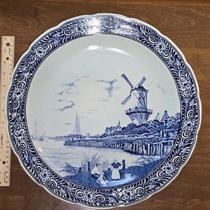 Könnte beinhalten: Ein großer runder Keramikteller mit gewelltem Rand, verziert mit einem blau-weißen Motiv einer niederländischen Windmühle, einem Hafen mit Segelbooten und Figuren im Vordergrund. Ein Lineal zeigt eine Breite von etwa 28 cm an.