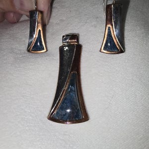 May include: A jewelry set consisting of a pendant and two matching earrings. Each piece features a flared, trapezoidal shape with a polished silver-tone metal finish, copper-colored accents, and a triangular inlay of dark blue textured stone.