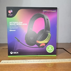 May include: A black PDP Afterglow Wave Wireless stereo gaming headset box featuring color-changing RGB lighting, a microphone, and Xbox branding. A ruler placed in front shows the box width is approximately 12 inches across the bottom edge.