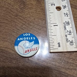 May include: A vintage Los Angeles Angels baseball pin. The pin is round with a blue and white design featuring a baseball with wings and the team name. A wooden ruler with inch measurements is next to the pin.