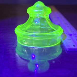 May include: A glowing, neon green glass jar with a lid. The jar has a rounded base and a decorative, tiered lid. The glass appears to be translucent, and the jar is reflecting on the surface below. A ruler is partially visible in the background.