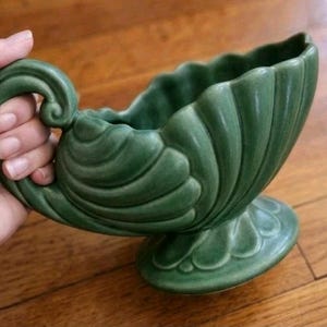 May include: Green ceramic planter with a handle and a scalloped rim. The planter has a pedestal base and a textured, shell-like design. The handle features a decorative swirl. The planter is a muted green color.