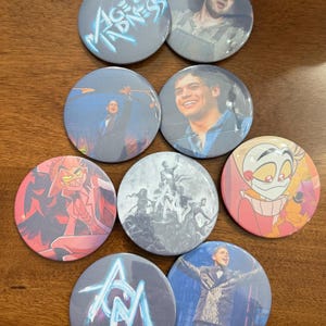 Könnte beinhalten: Eine Sammlung von neun runden Buttons mit verschiedenen Designs. Einige Buttons zeigen Bilder von Personen, während andere Text wie "Age of Madness" und "AM" im Neonstil anzeigen. Die Buttons liegen auf einer Holzoberfläche.