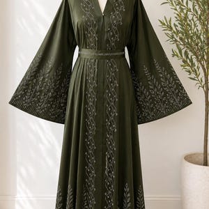 Puede incluir: Un vestido verde oliva con mangas largas y anchas y cuello en V. El vestido presenta intrincados bordados plateados en la parte delantera, las mangas y la cintura. Un cintur&oacute;n a juego ci&ntilde;e la cintura. El vestido se exhibe en un maniqu&iacute;.