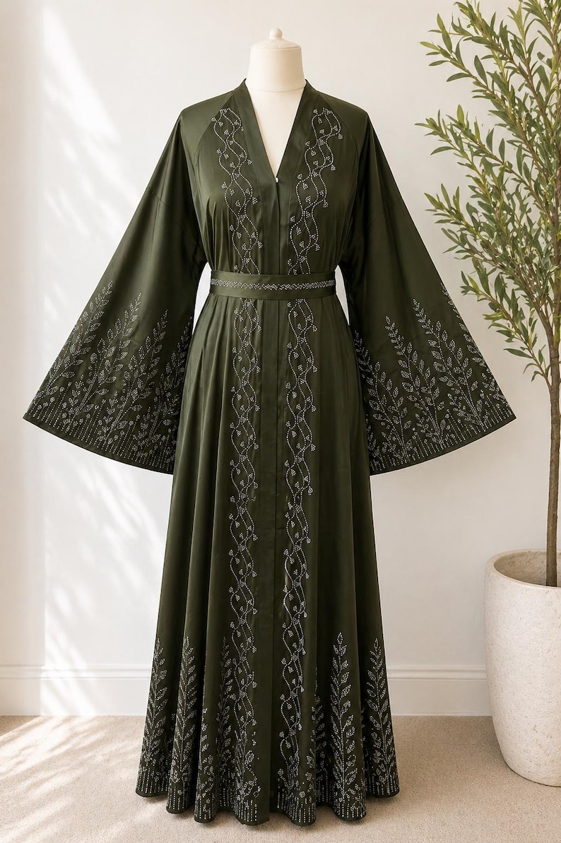 Puede incluir: Un vestido verde oliva con mangas largas y anchas y cuello en V. El vestido presenta intrincados bordados plateados en la parte delantera, las mangas y la cintura. Un cintur&oacute;n a juego ci&ntilde;e la cintura. El vestido se exhibe en un maniqu&iacute;.