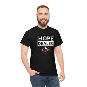 Puede incluir: Camiseta negra con el texto "HOPE DEALER" en un rectángulo blanco. Debajo, un círculo rojo con el contorno de Texas y las letras "H" y "C". La camiseta la lleva una persona.