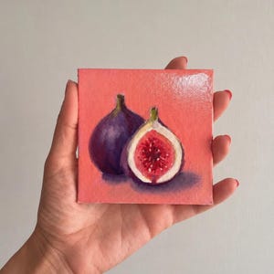 Peut inclure: Une petite peinture sur toile carrée représentant deux figues sur un fond rose corail uni. Une figue entière violet foncé est placée à côté d'une figue coupée en deux révélant un intérieur rouge vif avec des pépins et une peau blanche.