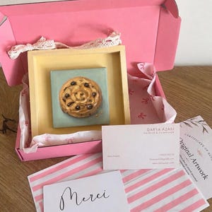 Peut inclure: Une petite peinture carrée représentant un cookie aux pépites de chocolat dans un cadre jaune clair, placée dans une boîte cadeau rose avec du papier de soie blanc. Inclut une carte de visite Darya Azari, une carte Merci et un certificat.