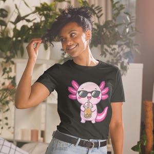 Könnte beinhalten: Ein schwarzes T-Shirt mit einer Grafik eines rosa Axolotls, das eine schwarze Sonnenbrille trägt und einen Plastikbecher mit braunem Bubble Tea mit einem rot-weiß gestreiften Strohhalm hält. Der Axolotl hat leuchtend rosa, federartige Kiemen.