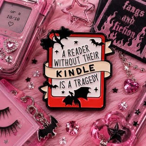 May include: A rectangular enamel pin with a white center and red border, featuring black bat silhouettes and a beige banner. The text reads: A READER WITHOUT THEIR KINDLE IS A TRAGEDY. The design includes small black stars and decorative accents.