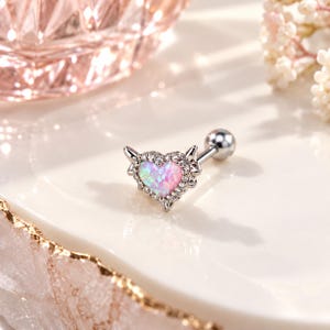 May include: A silver-toned barbell body jewelry piece featuring a heart-shaped pink opal stone at the center. The heart is framed by a halo of small clear crystals and accented with two small silver wings on the sides, attached to a metal post.