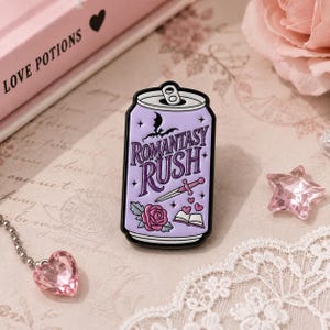 May include: A lavender-colored enamel pin shaped like a soda can with black outlines. The text ROMANTASY RUSH is printed in purple. Graphics include a black bat silhouette, a pink dagger, a pink rose, a small open book, and floating hearts.
