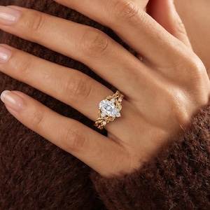 Può includere: Un anello in oro con una pietra preziosa bianca centrale a taglio ovale, affiancata da pietre bianche rotonde più piccole. La fascia presenta un design strutturato a forma di tralcio di vite che avvolge il dito, con finitura lucida.