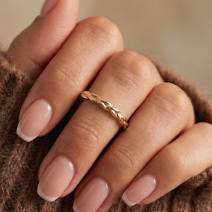 Puede incluir: Un anillo dorado con un diseño trenzado. El anillo se lleva en un dedo, con la mano apoyada en un suéter marrón. El patrón detallado del anillo es visible, destacando su artesanía. Joyería.