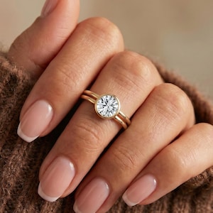 May include: A gold ring featuring a round, brilliant-cut clear gemstone set in a smooth bezel setting. The band is thin and polished, showcasing a minimalist design that highlights the central stone. The ring is worn on a finger with a neutral manicure.