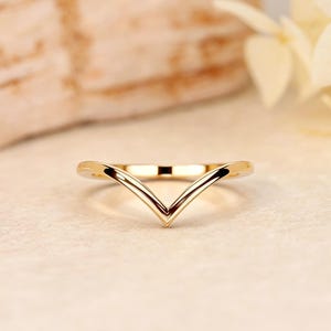 Puede incluir: Un anillo dorado en forma de V. El anillo tiene una superficie lisa y pulida y un diseño minimalista. El anillo se encuentra sobre un fondo neutro, lo que resalta su elegancia. Es probable que el anillo esté hecho de oro o metal dorado.