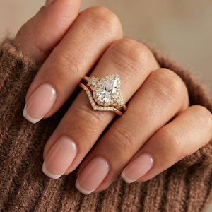 May include: A gold engagement ring set featuring a pear-shaped diamond center stone, surrounded by smaller diamonds. The set includes a curved band with additional diamond accents. The rings are displayed on a hand with a brown sweater.