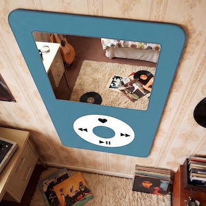 May include: A blue wall mirror shaped like a music player, with a white control panel featuring play, pause, and skip buttons. The mirror reflects a room with a guitar, records, and a fluffy rug, creating a retro music theme.