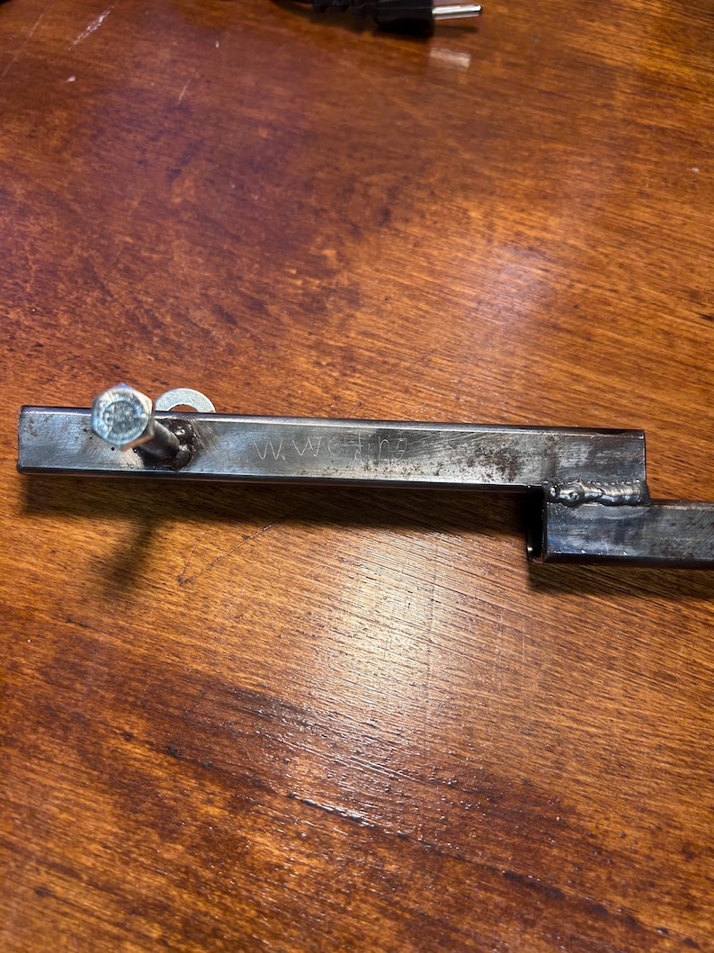 May include: A metal bracket with a bolt and washer attached. The bracket has the letters "WWE" stamped on it. The bracket is made of steel and has a welded joint. The bracket is on a wooden surface.