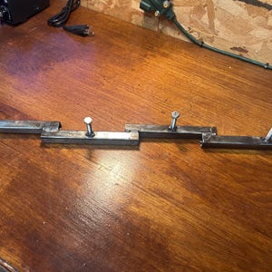 May include: Four metal brackets with bolts attached, arranged in a row. The brackets are made of rectangular steel tubing and are silver in color. The bolts are also silver. The brackets are on a wooden surface.