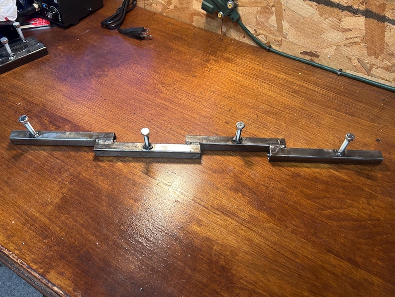 May include: Four metal brackets with bolts attached, arranged in a row. The brackets are made of rectangular steel tubing and are silver in color. The bolts are also silver. The brackets are on a wooden surface.